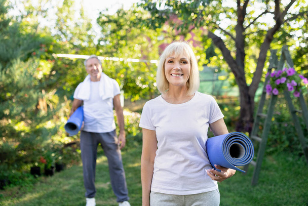 Healthy retirement lifestyle. Happy active senior couple holding yoga mats, looking to camera in sunny garden - Foto, immagini