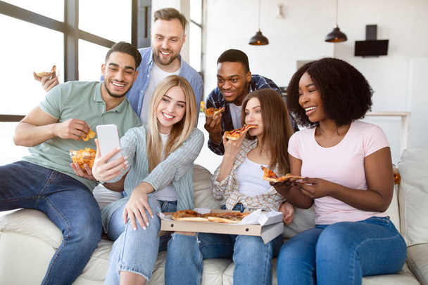 Gelukkig diverse vrienden hebben feest, zitten op de bank met pizza, chips en mobiele telefoon, socialiseren thuis - Foto, afbeelding