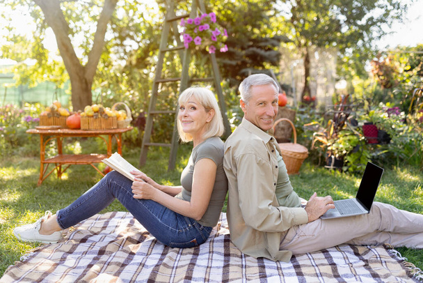 Happy senior couple resting on picnic blanket in garden, man using laptop computer, woman reading book - Photo, Image