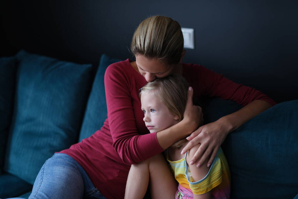 Woman and sad little daughter are sitting on couch and hugging - Foto, imagen
