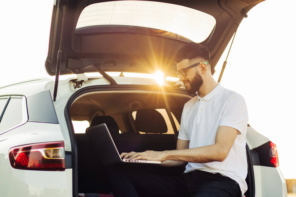 Young man in sunglasses enjoying a trip in his car, sitting in the trunk of a car with a laptop, Travel concept, summer vacation, vacation - Фото, зображення