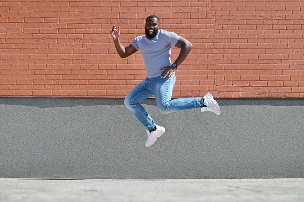 A dark-skinned man in a white tshirt and jeans jumping near the brick wall - Zdjęcie, obraz