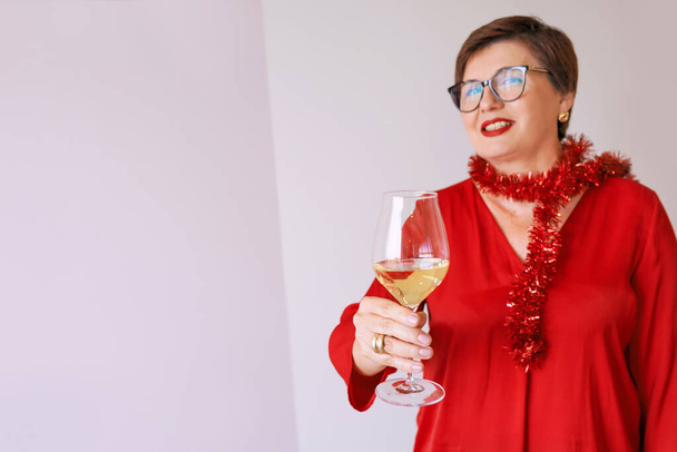 κομψή ώριμη ηλικιωμένη γυναίκα σε κόκκινη μπλούζα με ποτήρι λευκό κρασί γιορτάζει το νέο έτος. Διασκέδαση, πάρτι, στυλ, τρόπος ζωής, αλκοόλ, εορταστική έννοια - Φωτογραφία, εικόνα