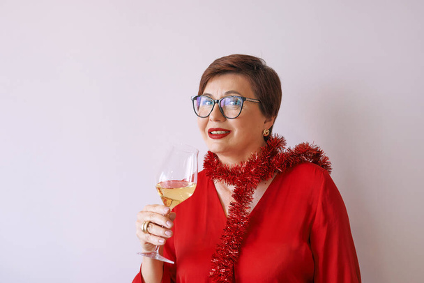新年を祝う白ワインのガラスと赤ブラウスでスタイリッシュな成熟したシニア女性。楽しい、パーティー、スタイル、ライフスタイル、アルコール、お祝いのコンセプト - 写真・画像