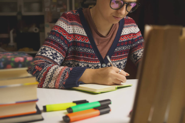 Молода щаслива жінка в окулярах вчиться вночі. Тисячолітній студент пише в блокноті. Розмиті книги та маркери на передньому плані. Освіта та концепція навчання. Тінту
. - Фото, зображення