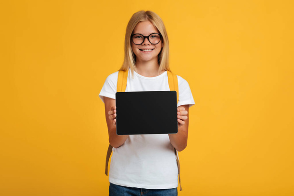 Mosolygós csinos tini szőke lány tanuló szemüvegben hátizsák mutatja tabletta üres képernyőn - Fotó, kép