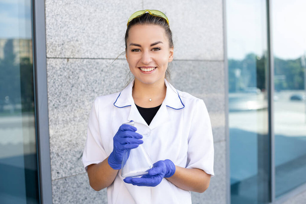 Chemistry, biology and medicine concept. A European happy female scientist in a medical gown and rubber gloves holding a chemical flask looking into the camera outside in front of a building - Foto, Imagen
