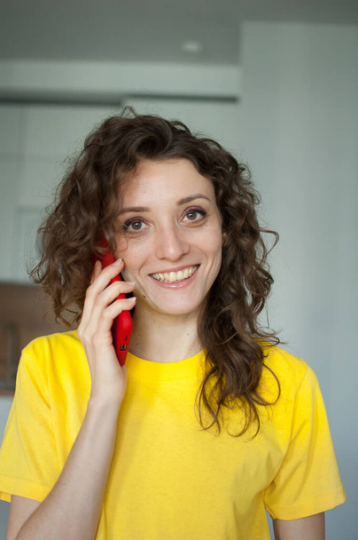 Menina encaracolado bonita em camiseta amarela está falando com alguém usando smartphone vermelho em casa em seu apartamento, posando com sorriso, conceito de pessoas felizes - Foto, Imagem