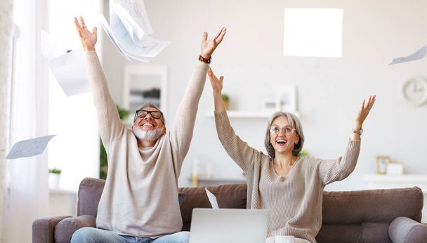 Podekscytowana szczęśliwa para seniorów siedząca na kanapie i rzucająca dokumentami papierowymi podczas płacenia rachunków online na laptopie w domu, emerytowany mąż i żona świętujący zatwierdzony kredyt hipoteczny - Zdjęcie, obraz