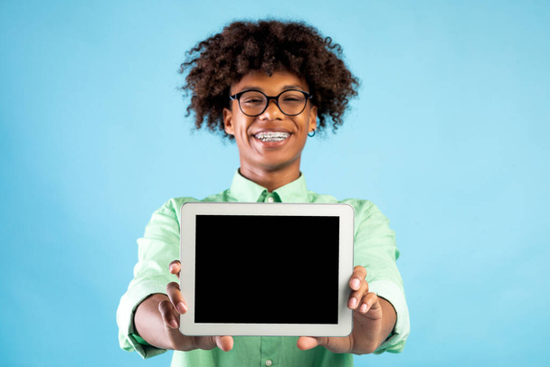 Smiling african american teen guy showing digital tablet with blank screen over blue studio background, copy space - Fotó, kép
