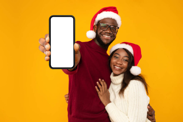 Schwarzes Paar in Weihnachtsmützen mit Handybildschirm, gelbem Hintergrund - Foto, Bild