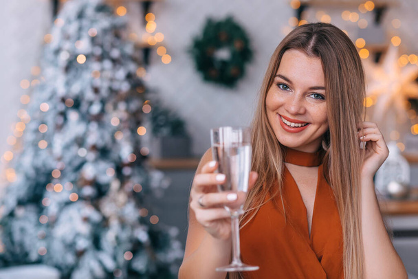 Kobieta w bluzce stoi na tle choinki w oczekiwaniu na przyjęcie świąteczne, trzymając kieliszek szampana w dłoni. - Zdjęcie, obraz