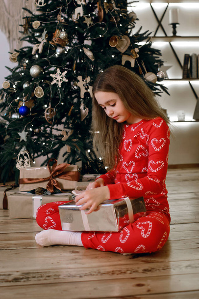 Lapsi tyttö punaisessa pyjamassa istuu koristellun joulukuusen lähellä ja avaa joululahjan. Tunnelma loma ja onnellinen lapsuus pystysuora valokuvaus. Talvi joulu lomat - Valokuva, kuva