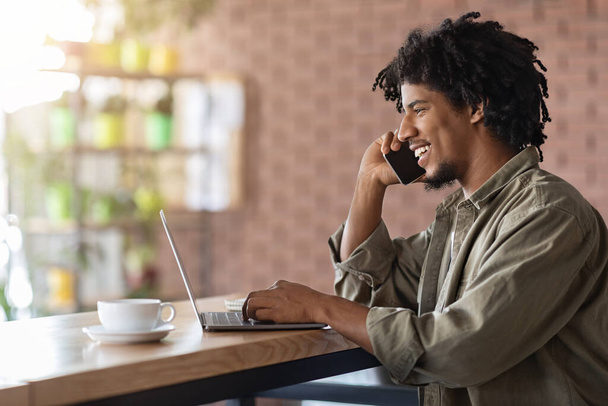 Kafede dizüstü bilgisayarla çalışırken cep telefonuyla konuşan siyahi serbest çalışan adam. - Fotoğraf, Görsel