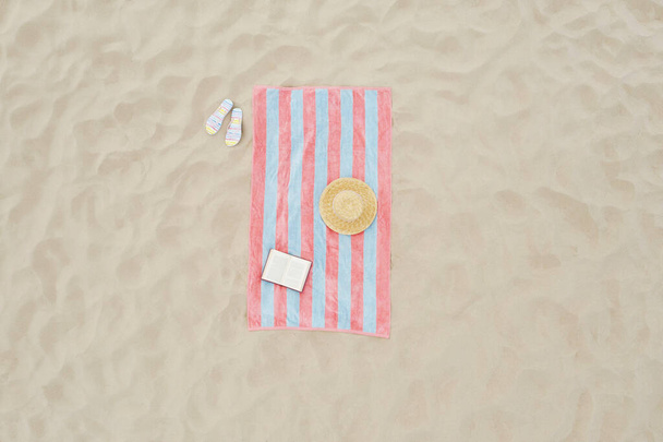 Striped beach towel, book, straw hat and flip flops on sand, aerial view - Photo, Image