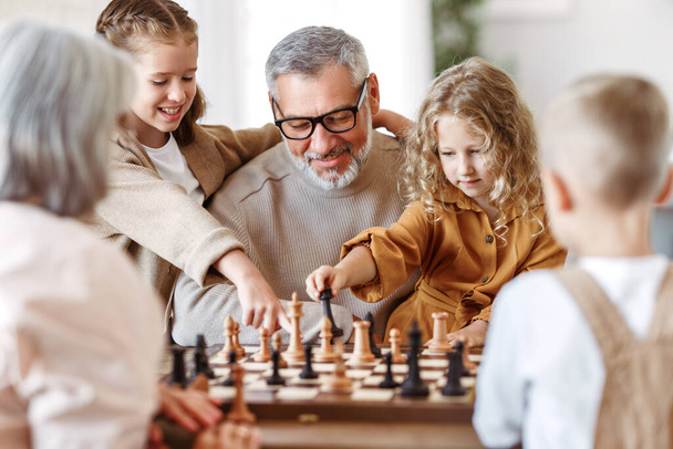Joyful children brother and sister playing chess while sitting in living room with senior grandparents while spending time together on weekend, kids sitting at table with chessboard and smiling - Foto, imagen
