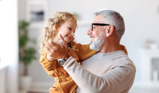 Elegante liefdevolle zorgzame grootvader die kijkt naar zijn schattige kleindochter, schattig meisje en positieve opa die hand in hand danst in de woonkamer thuis. Gezinsconcept - Foto, afbeelding