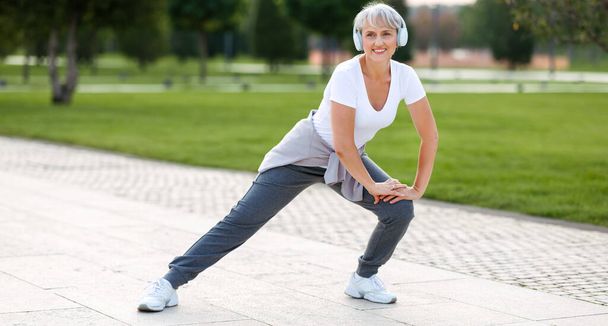 Šťastná pozitivní zralé sportovkyně při venkovním cvičení, starší žena nosí sluchátka a sportovní oblečení zahřívá svaly, dělá boční dřepy na jedné noze, těší aktivní životní styl venku v parku - Fotografie, Obrázek