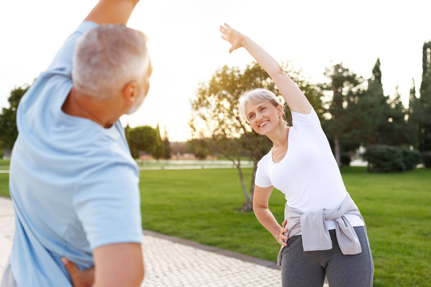 Healthy mind and body. Full length shot of happy smiling mature family man and woman in sportswear stretching arms while warming up together outdoors in park on sunny morning. Active lifestyle - Foto, Imagem