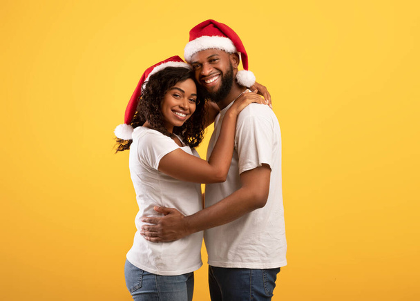 Afrikaanse amerikaanse minnaars in Santa hoeden omarmen op gele studio achtergrond, vieren Nieuwjaar 2022 of Kerstmis - Foto, afbeelding
