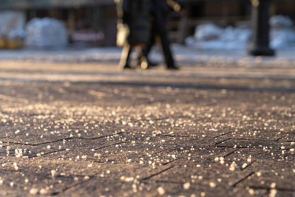 Az úton lévő jégmentesítő kémiai reagens télen biztonságossá teszi az utat. A járda tele van technikai sóval, az előtérre koncentrál, a homályos hátterű emberekre.. - Fotó, kép