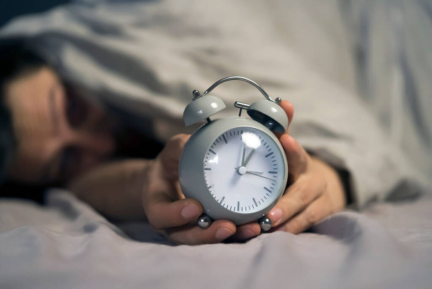 Egy fiatalember keze a takaró alól egy retro vintage ébresztőórát tart.. - Fotó, kép