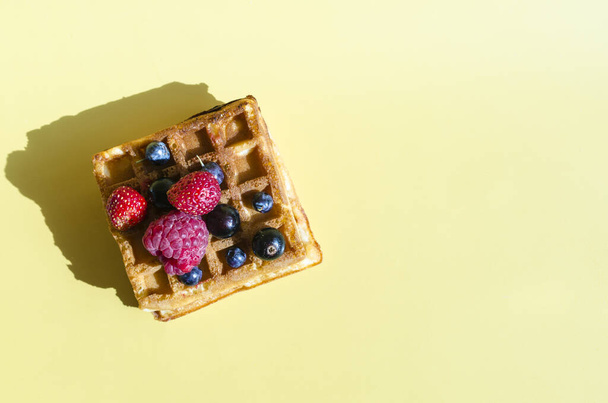 A waffle with different berries of black and red color on a yellow background is hard light. Homemade cakes crispy waffles on a colored background top view of kopi space - Foto, afbeelding