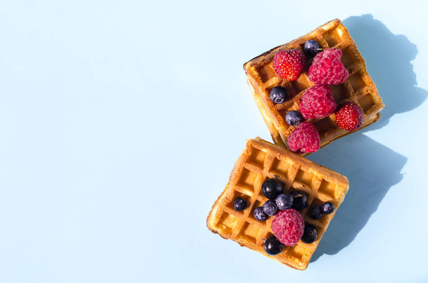Waffles with black and red berries on a blue background of hard light. Homemade cakes crispy waffles on a colored background top view - Foto, Imagem