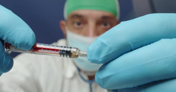 Widok lekarza w białym fartuchu, masce chirurgicznej, niebieskich rękawiczkach i nakryciu głowy za pomocą strzykawki podczas badania laboratoryjnego szczepionek koronawirusowych - Materiał filmowy, wideo