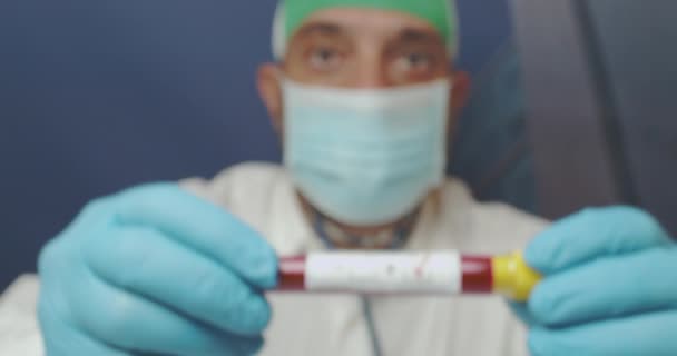 解析室の医師の手は、ビデオカメラで患者のcovid19陽性血液検査の結果を示しています.試験結果は試験管に書かれ、医師は白いコートと青い手袋を着用します。 - 映像、動画