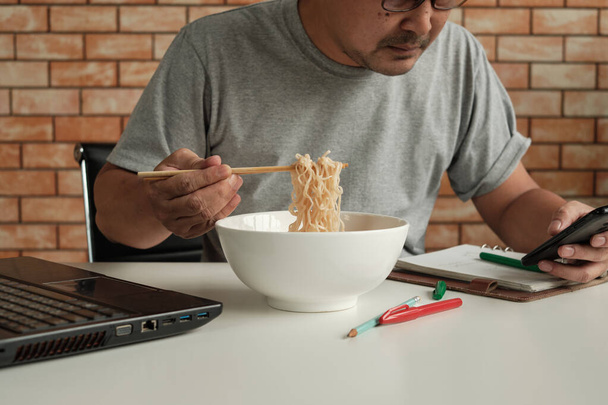 Férfi dolgozó elfoglalt dolgozik egy laptop és a mobil telefon, használja evőpálcikát enni instant tészta alatt munkahelyi ebédidőben, mert gyors, ízletes, és olcsó. Idővel ázsiai gyorsétterem, egészségtelen életmód. - Fotó, kép