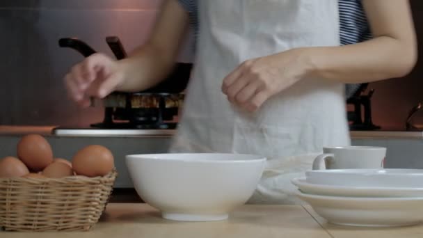 Una cuoca in grembiule bianco sta incrinando un uovo in una tazza per preparare un pasto su un tavolo di legno nella cucina di casa. Mangiare tuorli d'uovo è una colazione sana. - Filmati, video