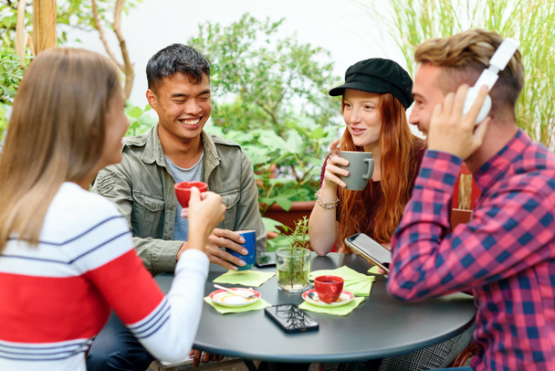 Group of diverse young students enjoying a relaxing break seated around an outdoor table laughing and joking while enjoying a mug of coffee - Photo, Image