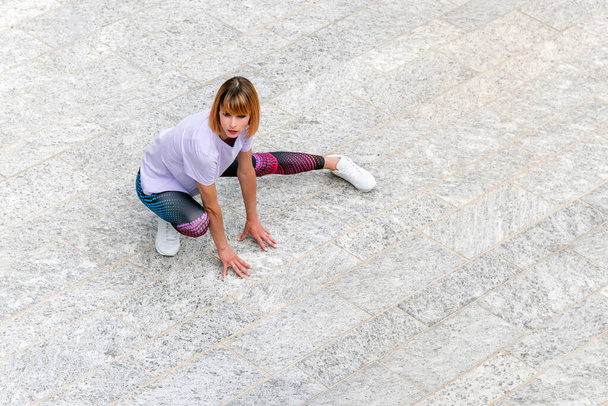 Du haut du corps complet de la jeune femme en forme dans des vêtements de sport élégants et des baskets faisant des exercices d'étirement pour les jambes sur la place urbaine pavée pendant l'entraînement de fitness en plein air - Photo, image