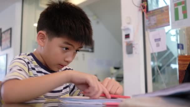 Schöner Junge macht Hausaufgaben, während er mit rotem und grünem Bleistift in der Lobby des Familienunternehmens zeichnet - Filmmaterial, Video