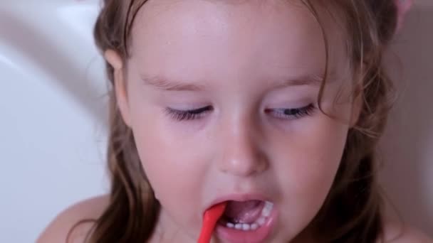Ένα κοριτσάκι 3 ετών βουρτσίζει τα δόντια της με μια κόκκινη οδοντόβουρτσα στο μπάνιο του σπιτιού. Υγειονομική περίθαλψη, οδοντιατρική υγιεινή, άνθρωποι και ομορφιά - Πλάνα, βίντεο