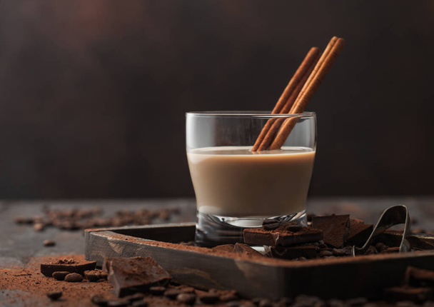 Irish cream baileys liqueur in crystal glass with cinnamon, coffee beans and powder with dark chocolate in wooden tray on dark wood background. Space for text - Foto, imagen