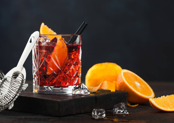 Negroni cocktail in crystal glass with orange slice and fresh raw oranges on chopping board with strainer on wooden background. - Photo, Image