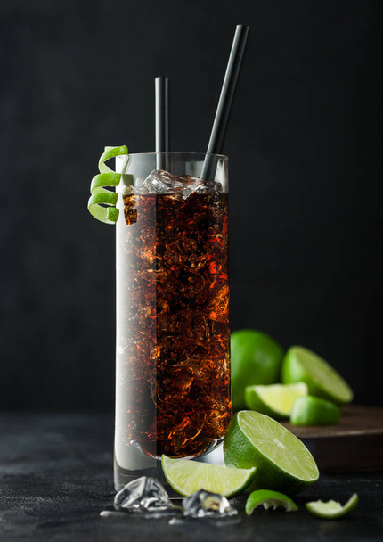 Cuba Libre cocktail in highball glass with ice and lime peel with straw and fresh limes on black table background. - Φωτογραφία, εικόνα