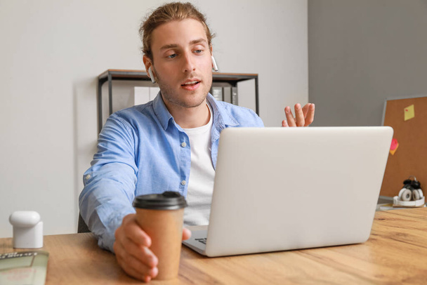 Handsome young man with cup of coffee video chatting in office - 写真・画像