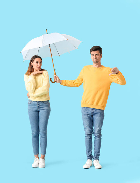 Displeased guy sharing umbrella with young woman on color background - 写真・画像