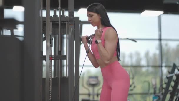 Een brunette in een roze pak trekt aan een touw in een crossover arm voor biceps training. Handtraining in een trainer. Professionele vrouwelijke instructeur - Video