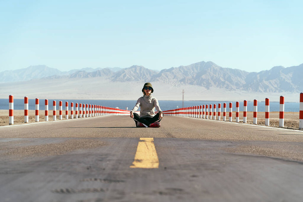 азіатська жінка турист, сидячи посеред порожньої відкритої дороги з озером і прокатними горами на задньому плані, нога схрещена
. - Фото, зображення