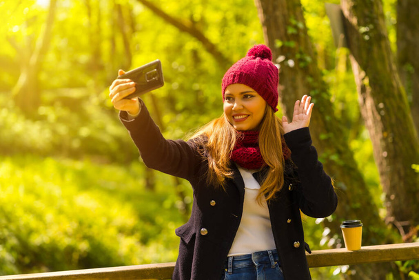Молодой человек в черной куртке, шарфе и красной шерстяной шляпе наслаждается в осеннем парке, машет на видео-звонок с телефона - Фото, изображение