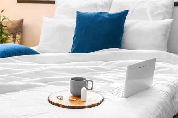 Δίσκος με κύπελλο και ακουστικά, φορητό υπολογιστή σε μαλακό κρεβάτι - Φωτογραφία, εικόνα