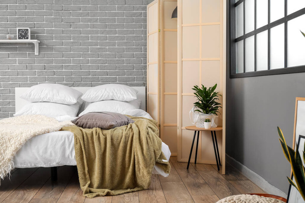 Cama confortável, tela dobrável e mesa com planta de sala perto da parede de tijolo cinza - Foto, Imagem
