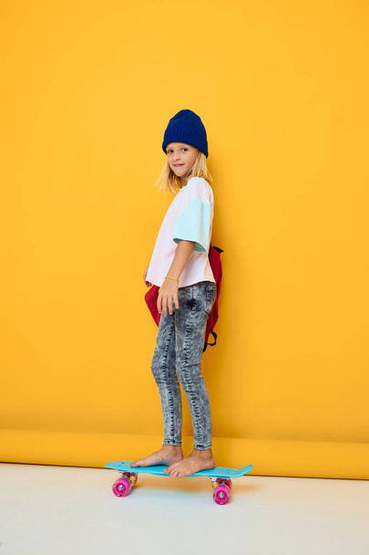 portret van een meisje gelukkig schattig kind met skateboard kid met rugzak gele achtergrond - Foto, afbeelding