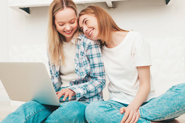 Two young beautiful smiling female in trendy summer clothes. Sexy carefree women posing in light interior in studio. Positive models having fun indoors.Using computer laptop.Looking at notebook screen - Foto, Imagem