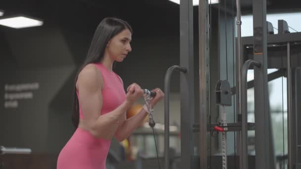 Una mujer morena con un traje rosa tira de una cuerda en un brazo cruzado para el entrenamiento del bíceps. Entrenamiento de manos en un entrenador. Instructora profesional - Imágenes, Vídeo