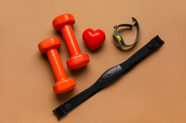 Fitness tracker, chest belt, dumbbells and red heart on color background - Photo, Image
